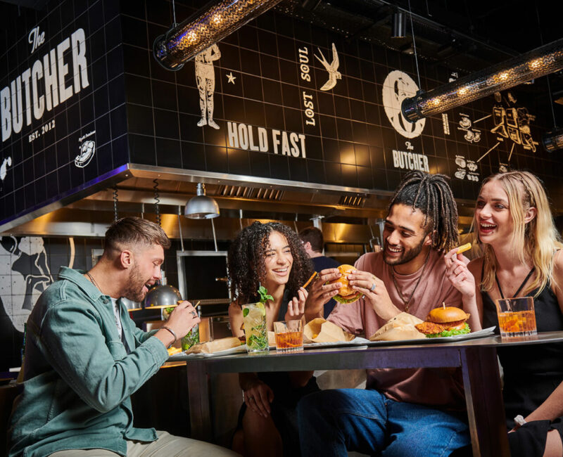 A lively group of four friends smiling and sat around a table with multiple pizzas.