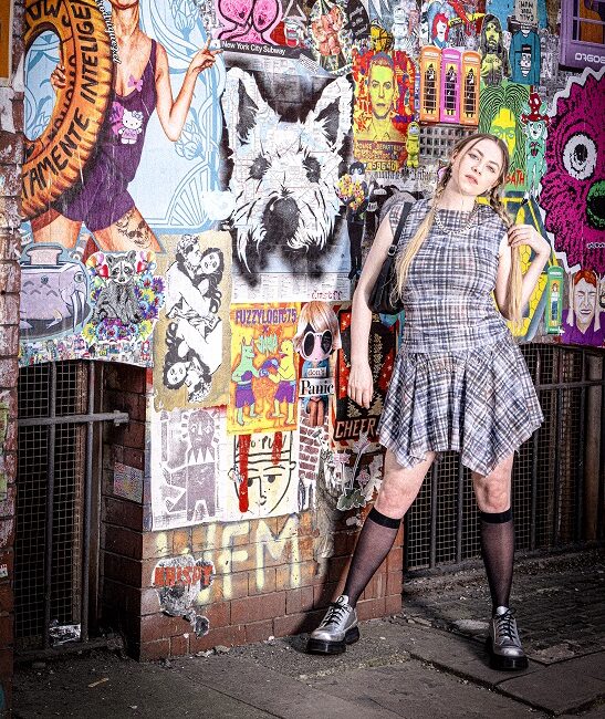 Girl wearing a check dress with knee high black socks. Background is full of art and graffiti.