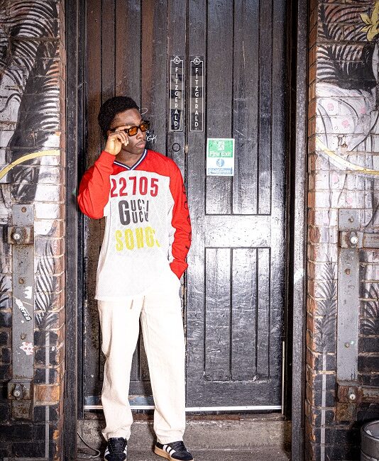 Man wearing American style red varsity top, leaning against a black door.