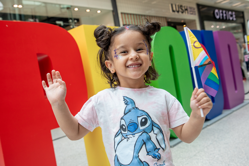 Little girl waving a LGBTQ+ flag in front of giant multi-coloured "PROUD" letters