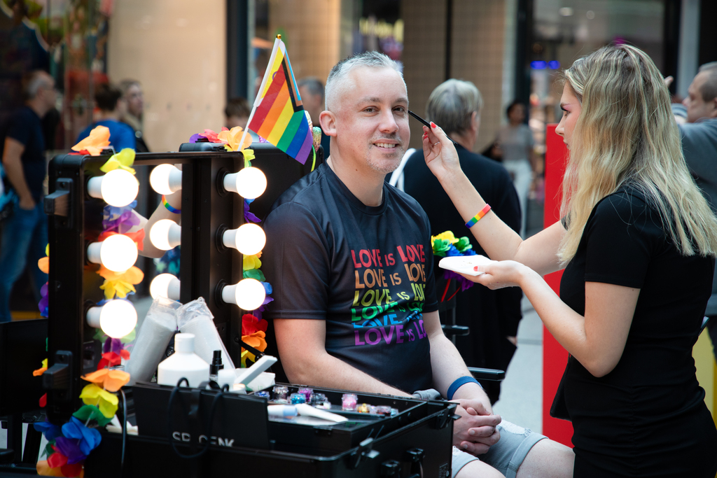 Man smiling at the camera whilst a make-up artist applies glitter to his face.