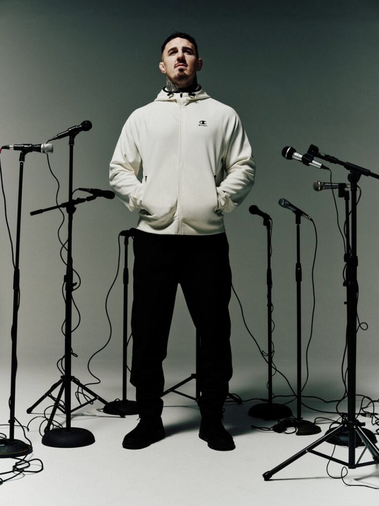Tom Aspinall posing with lots of mic stands surrounding him wearing grey CHAMPION hoodie